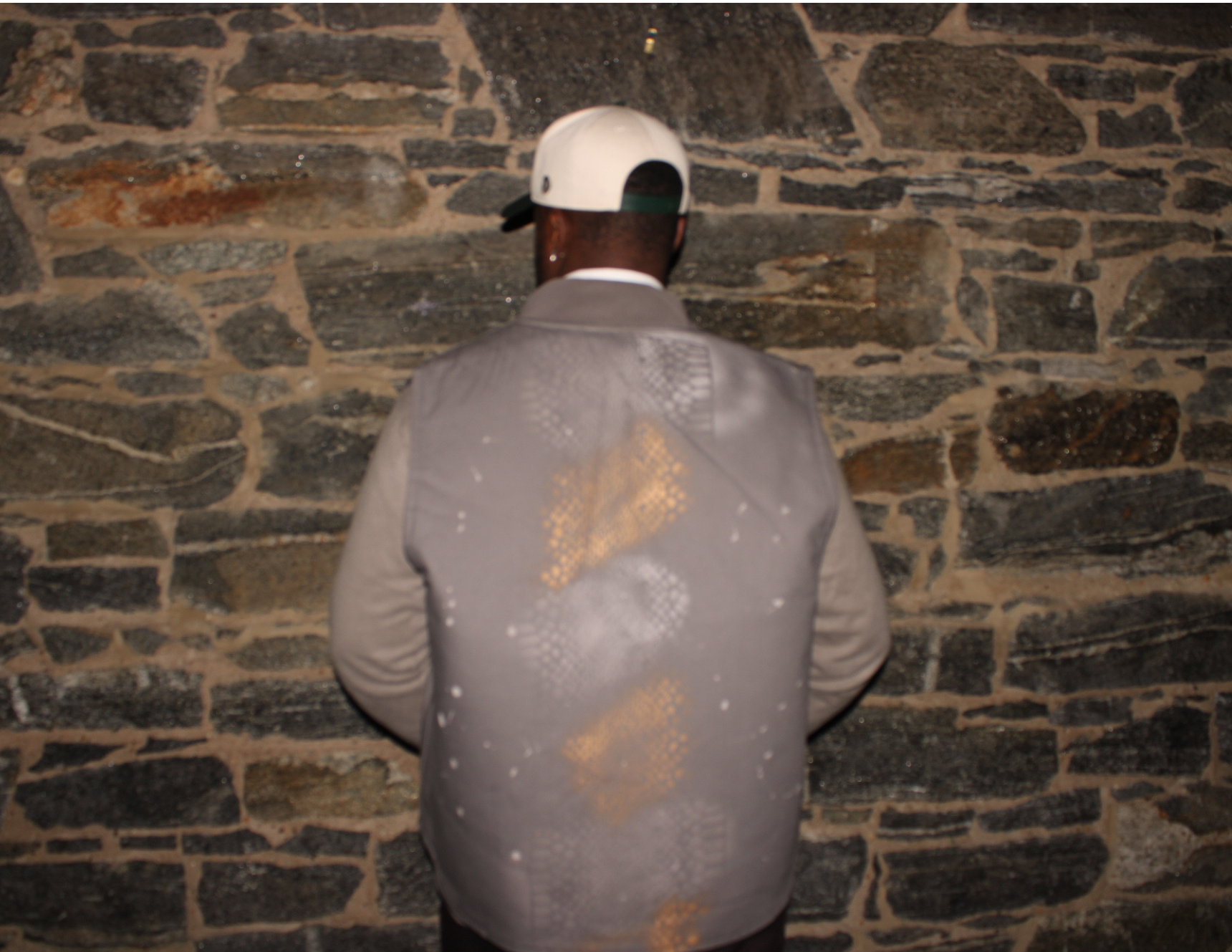 Person wearing a distressed gray jacket and white cap standing against a stone wall.